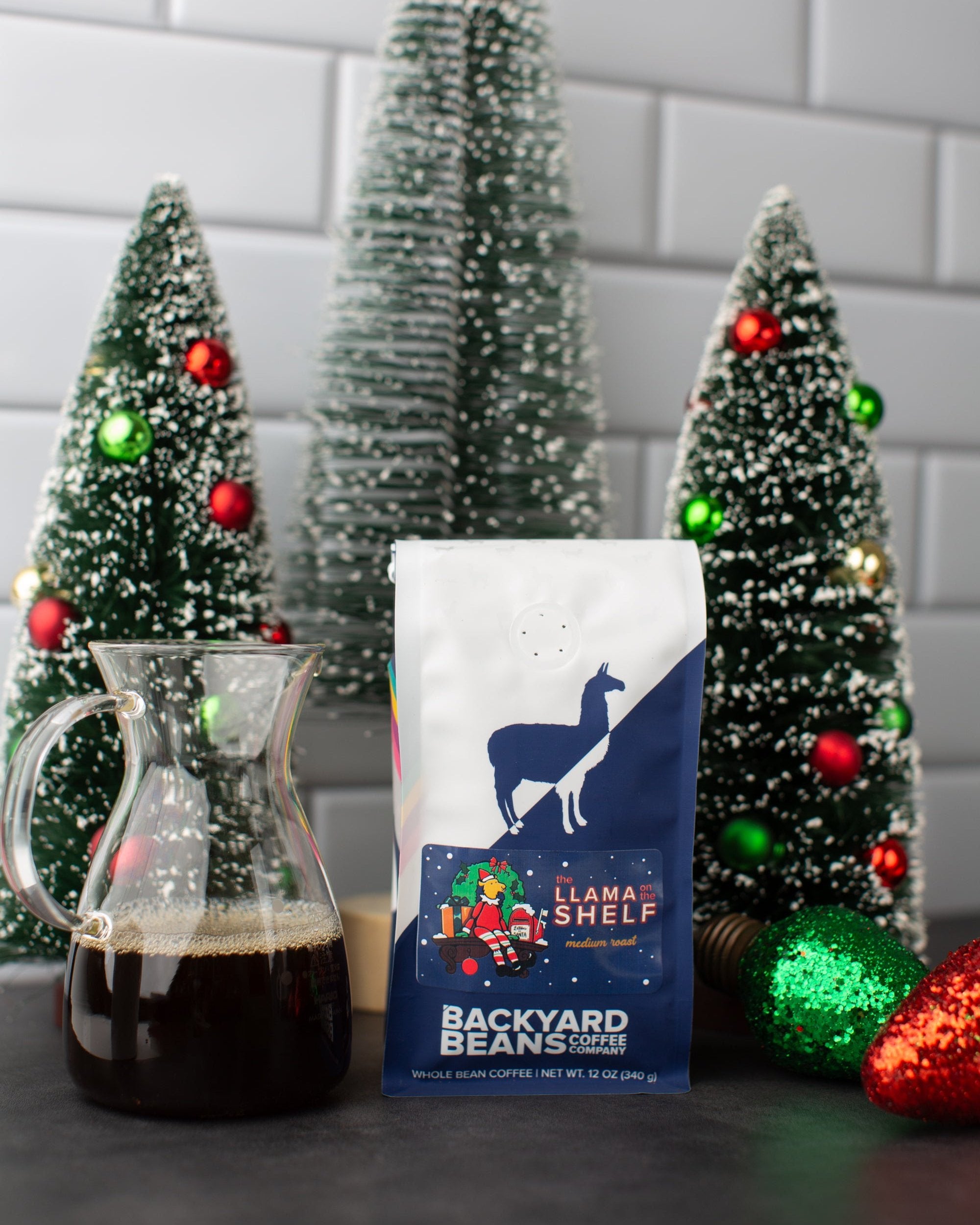 Image of Llama on the Shelf coffee roast next to coffee carafe and holiday trees in kitchen.