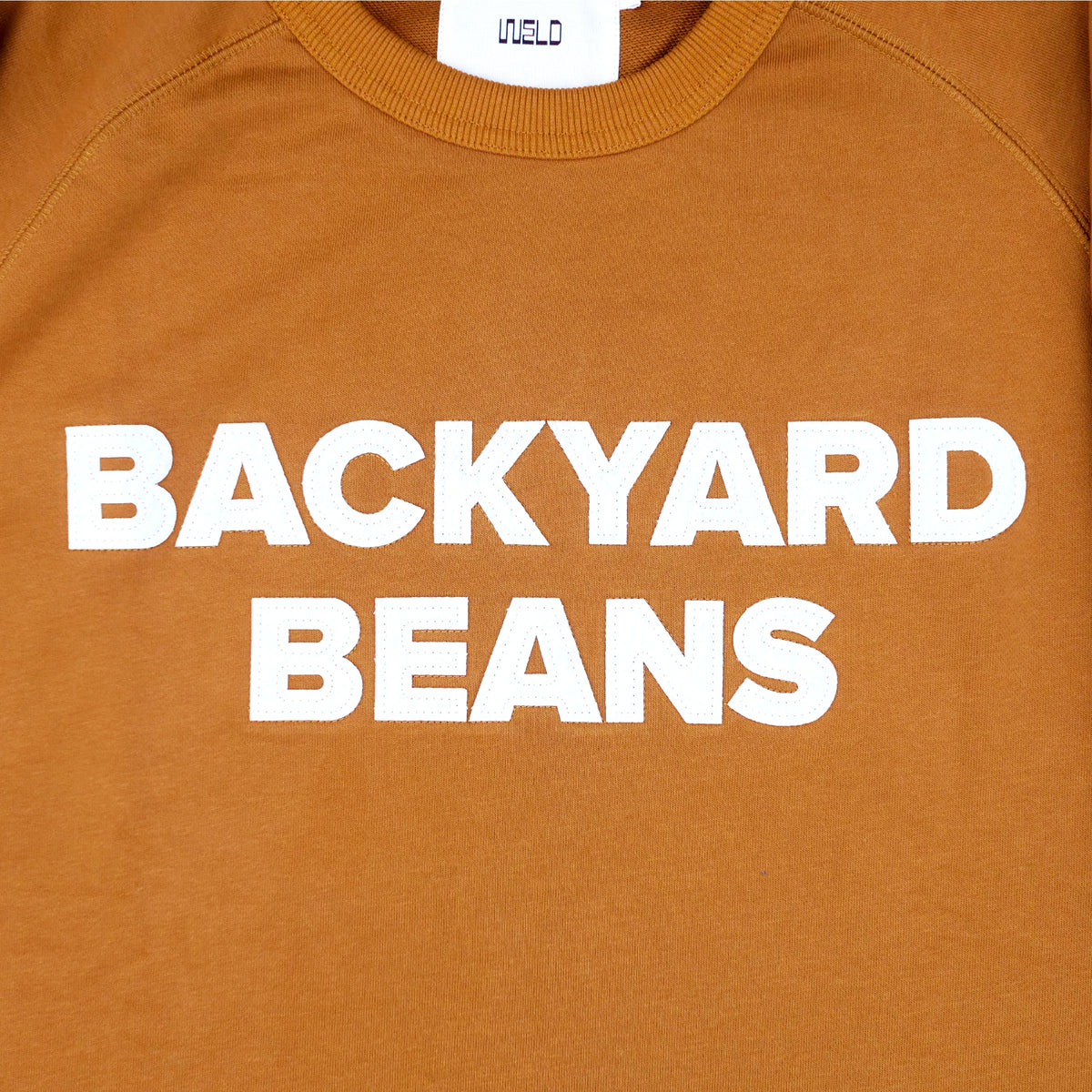 Close-up of a rust orange crewneck sweatshirt with 'BACKYARD BEANS' printed in bold white capital letters across the chest. The inner collar label shows the brand 'WELD'.