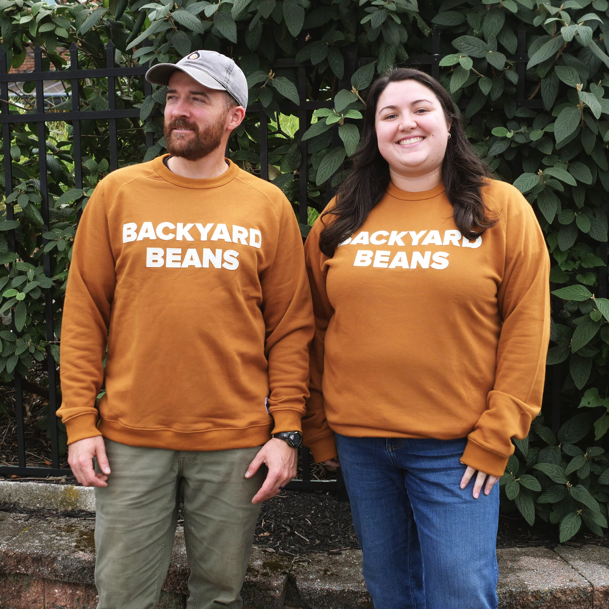 A man and a woman standing outdoors in front of green foliage, wearing matching rust orange sweatshirts with 'BACKYARD BEANS' printed in white across the chest.