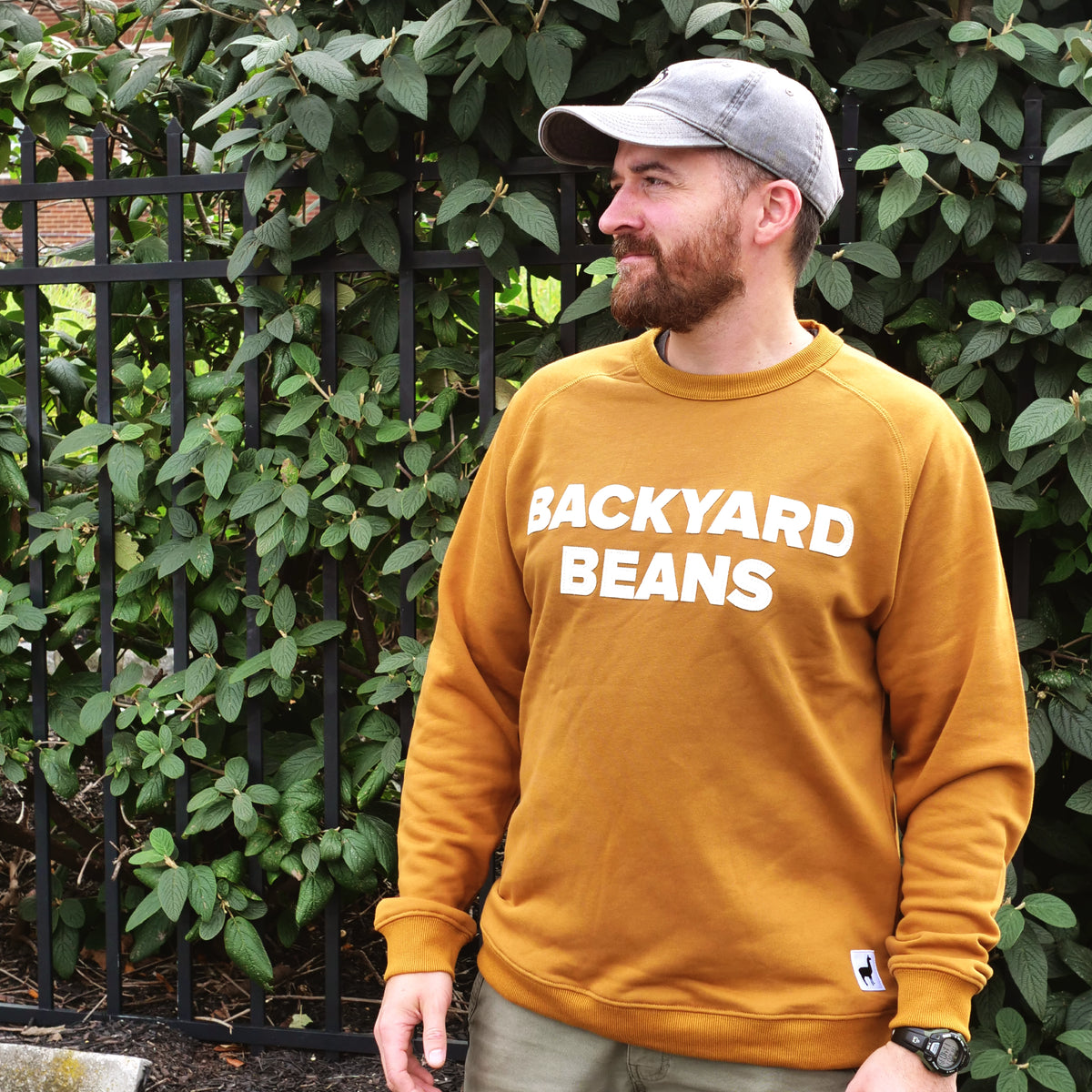 A man wearing a rust orange crewneck sweatshirt with 'BACKYARD BEANS' printed in white across the chest, standing outdoors in front of a black metal fence and green leafy bushes. He is also wearing a gray baseball cap and khaki pants.