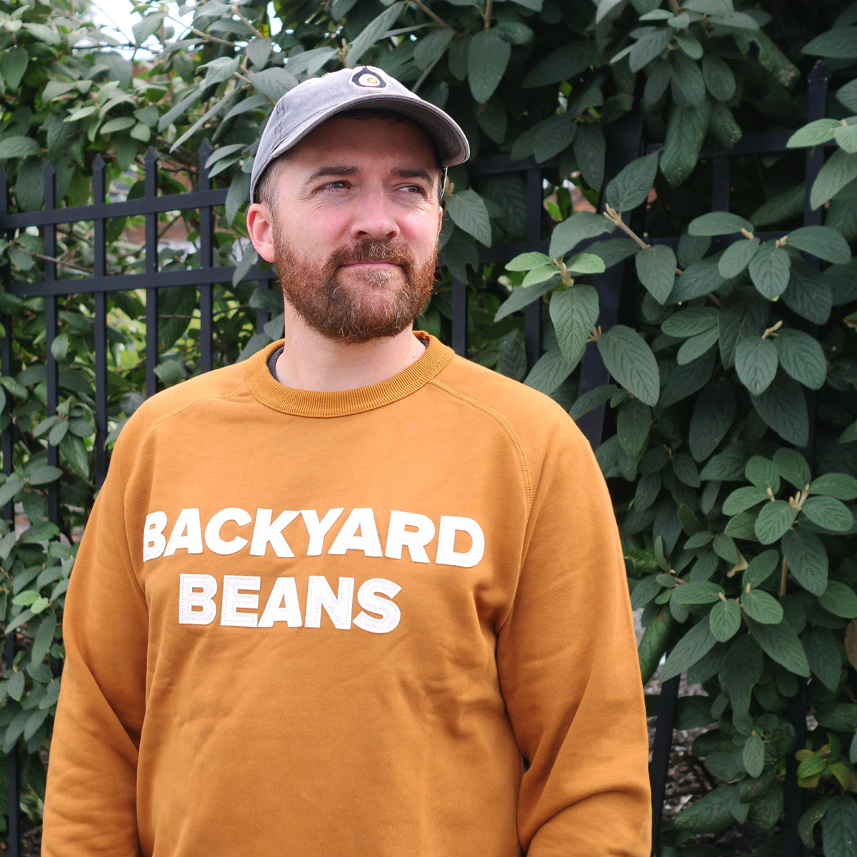 A man wearing a rust orange crewneck sweatshirt with 'BACKYARD BEANS' printed in bold white letters, paired with a gray baseball cap, standing outdoors in front of a black metal fence and green foliage.
