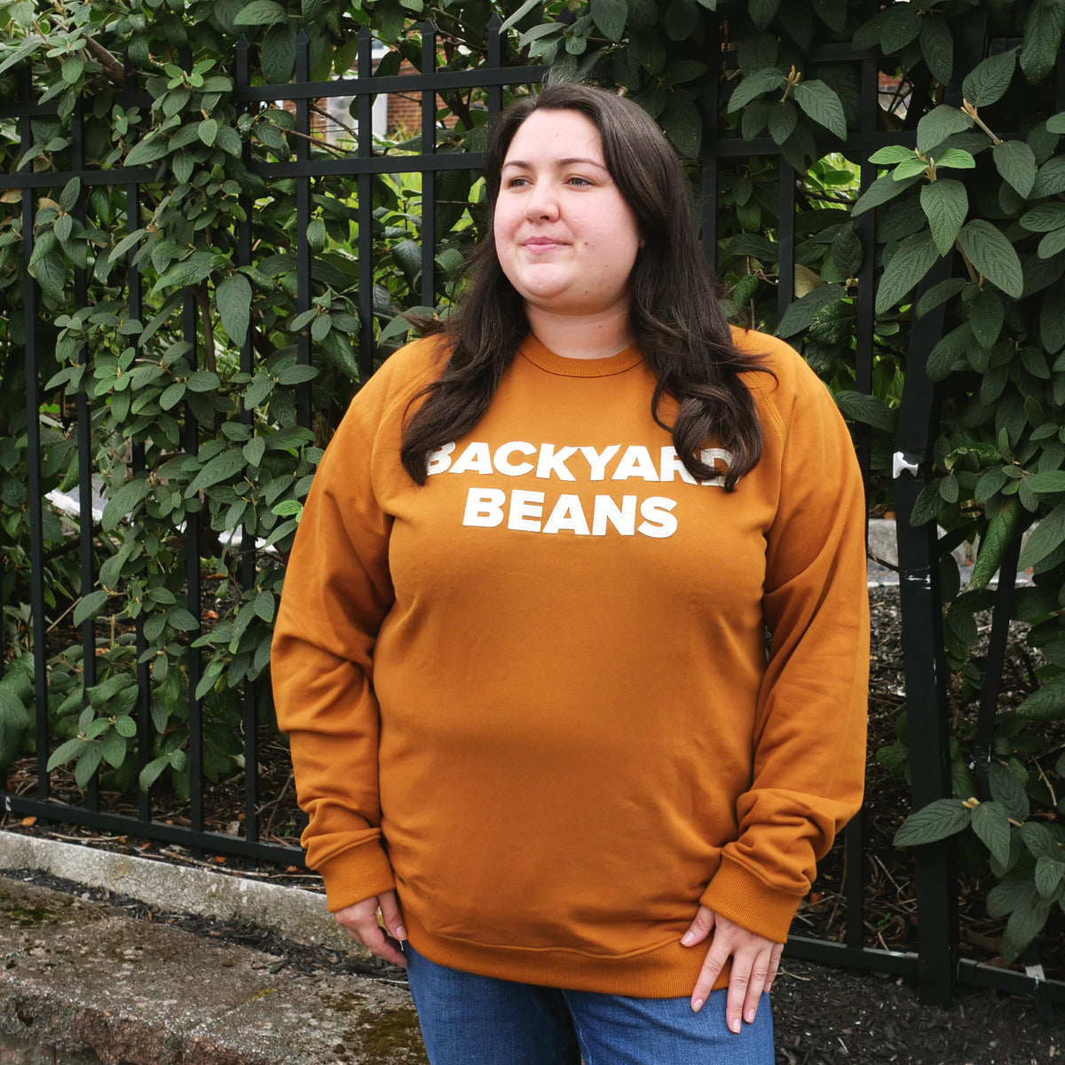 A woman wearing a rust orange crewneck sweatshirt with 'BACKYAK BEANS' printed in white across the chest, standing outdoors in front of a black metal fence and green ivy.