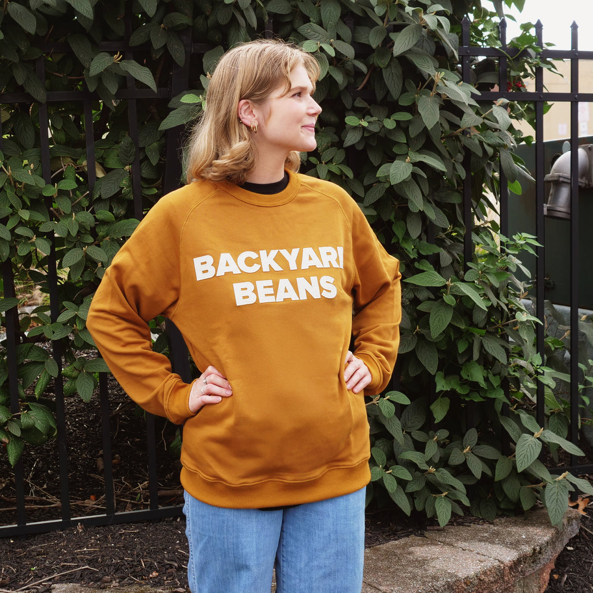 A woman wearing a rust orange sweatshirt with 'BACKYARI BEANS' printed in white across the chest, paired with blue jeans, standing outdoors in front of a black metal fence and green ivy.