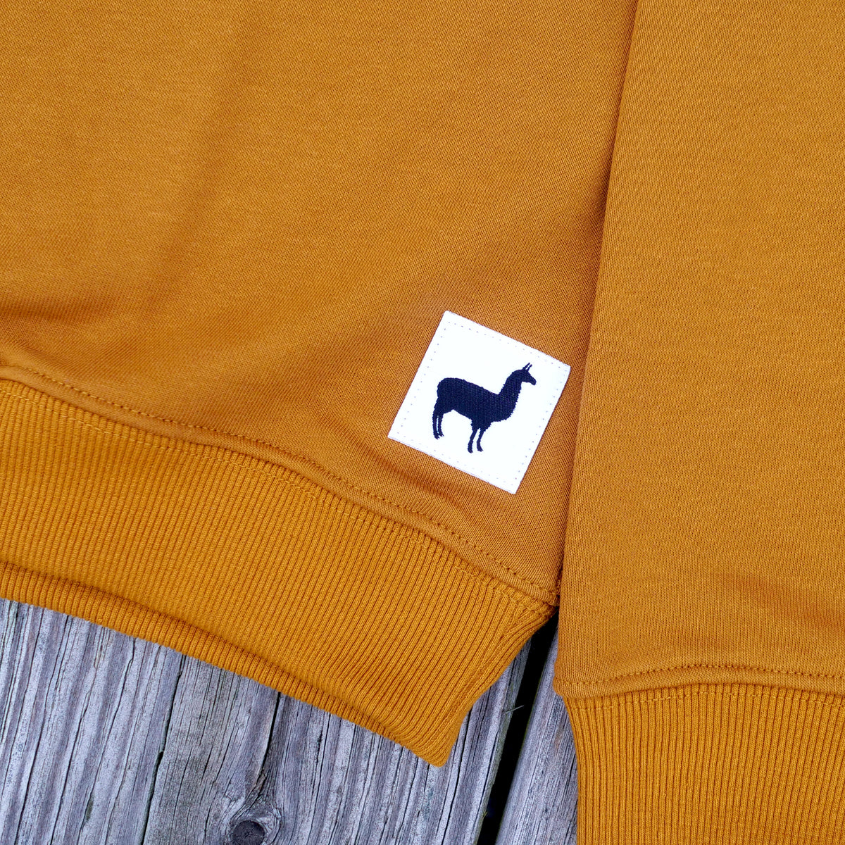 Close-up of a rust orange sweatshirt laid flat on a weathered gray wooden surface, with a small white square patch featuring a black llama silhouette near the cuff.