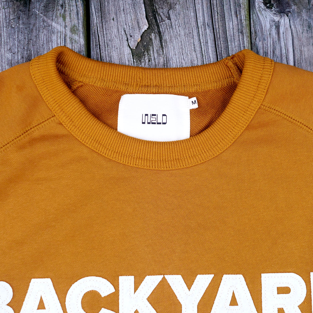 Close-up of rust orange crewneck sweatshirt with a white neck label reading 'WELD' and partial white text at the bottom, set against a weathered gray wood background.