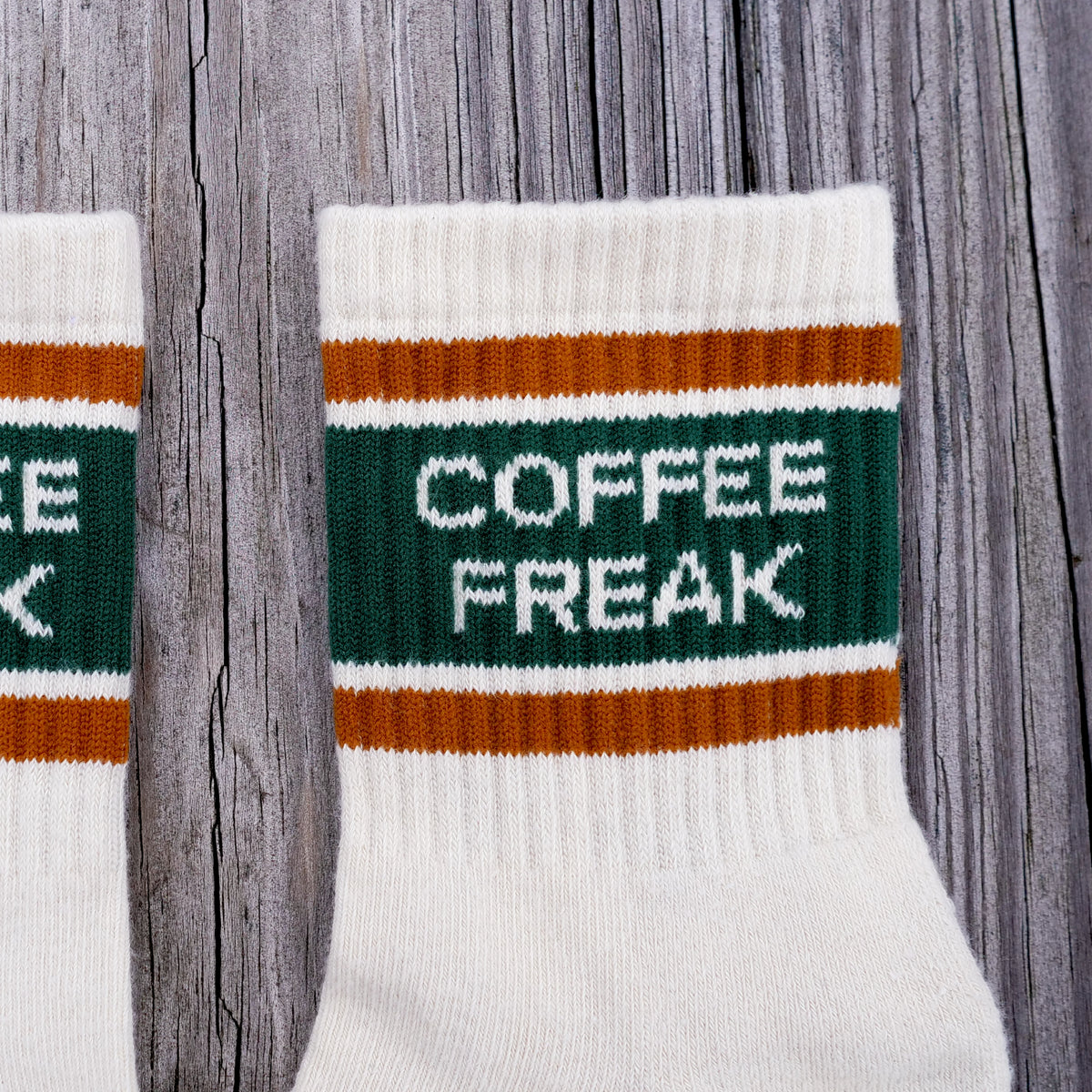 A close-up of a white knit crew sock with a dark green band reading 'COFFEE FREAK' in white letters, bordered by rust-orange stripes, laid on a weathered gray wood background.