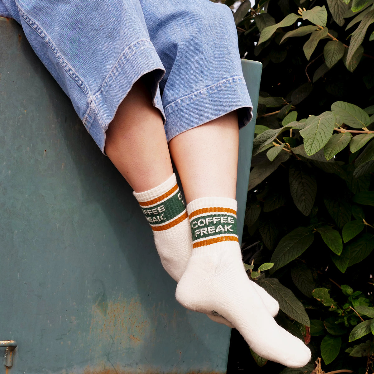 A person wearing white crew socks with green and orange stripes and the words 'COFFEE FREAK' sits on a teal metal surface with green foliage in the background.