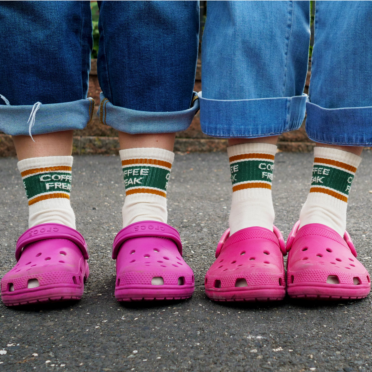 Two people standing outdoors wearing bright pink Croc-style clogs and white crew socks with green and orange stripes and 'COFFEE FREAK' text.
