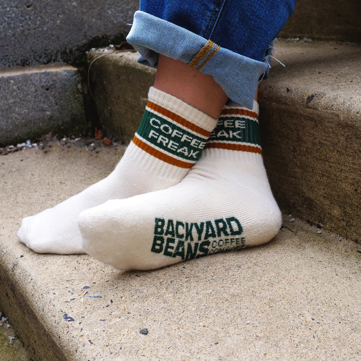 A person wearing off-white crew socks with green and orange stripes near the cuff, one sock reading 'COFFEE FREAK' and the other reading 'BACKYARD BEANS COFFEE COMPANY', shown on concrete steps.