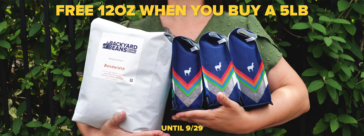 A person outdoors holding a large white bag of Backyard Beans Bandwidth beans next to three navy blue 12oz bags of coffee. Yellow promotional text at the top reads 'FREE 12OZ WHEN YOU BUY A 5LB' and 'UNTIL 9/29'.