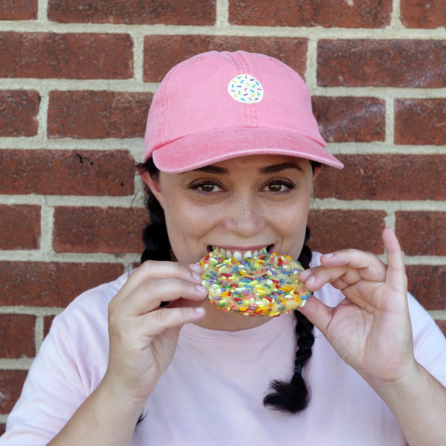Image of woman in coral dad hat with embroidered cookie hat biting a sprinkle sugar cookie